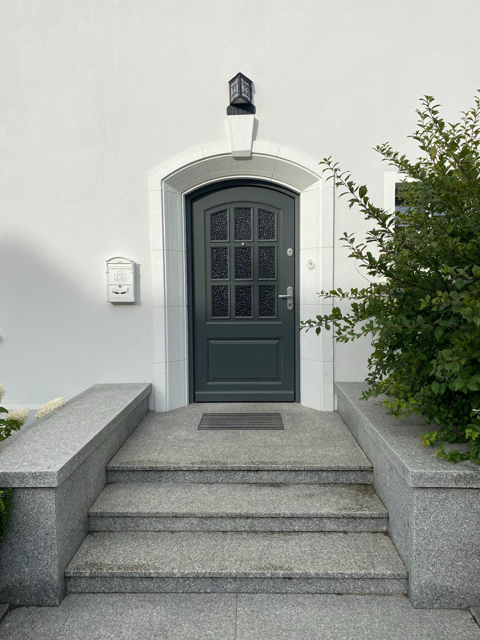 Eleganckie, grafitowe drzwi wejściowe z szybkami, osadzone w białej fasadzie z kamiennymi schodami i ozdobnym oświetleniem. Skrzynka pocztowa po lewej, zieleń po prawej.