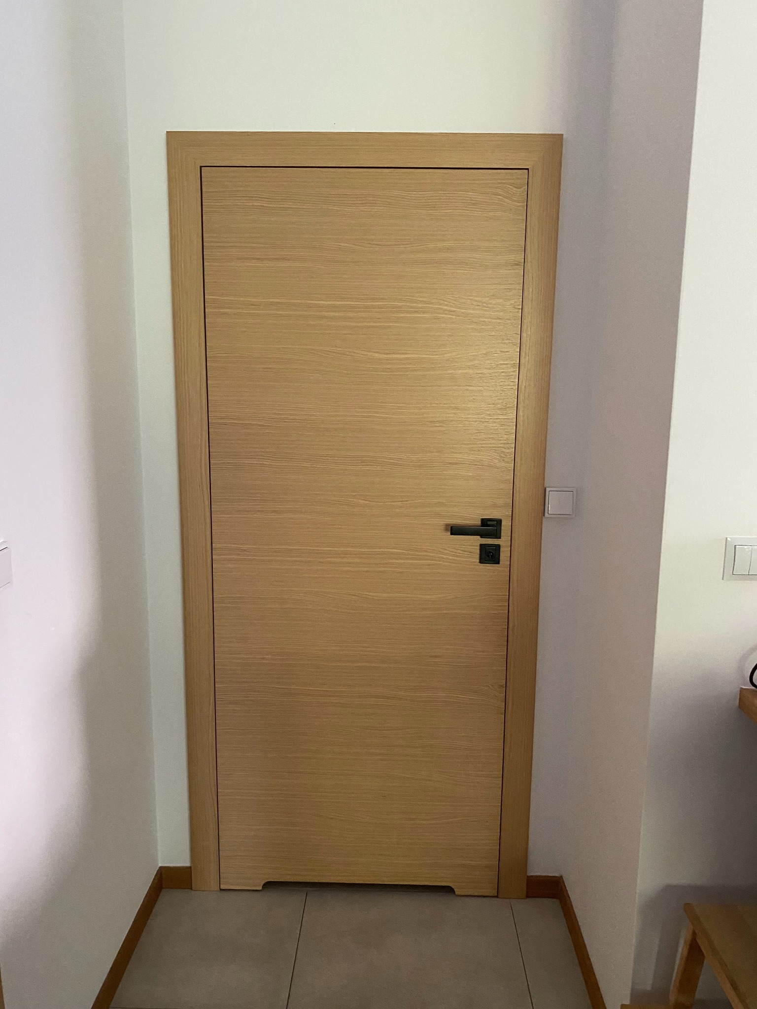 Jasne, drewniane drzwi wewnętrzne z czarną klamką, wstawione w białą ścianę. Widoczne szare płytki podłogowe i fragment listwy przypodłogowej.