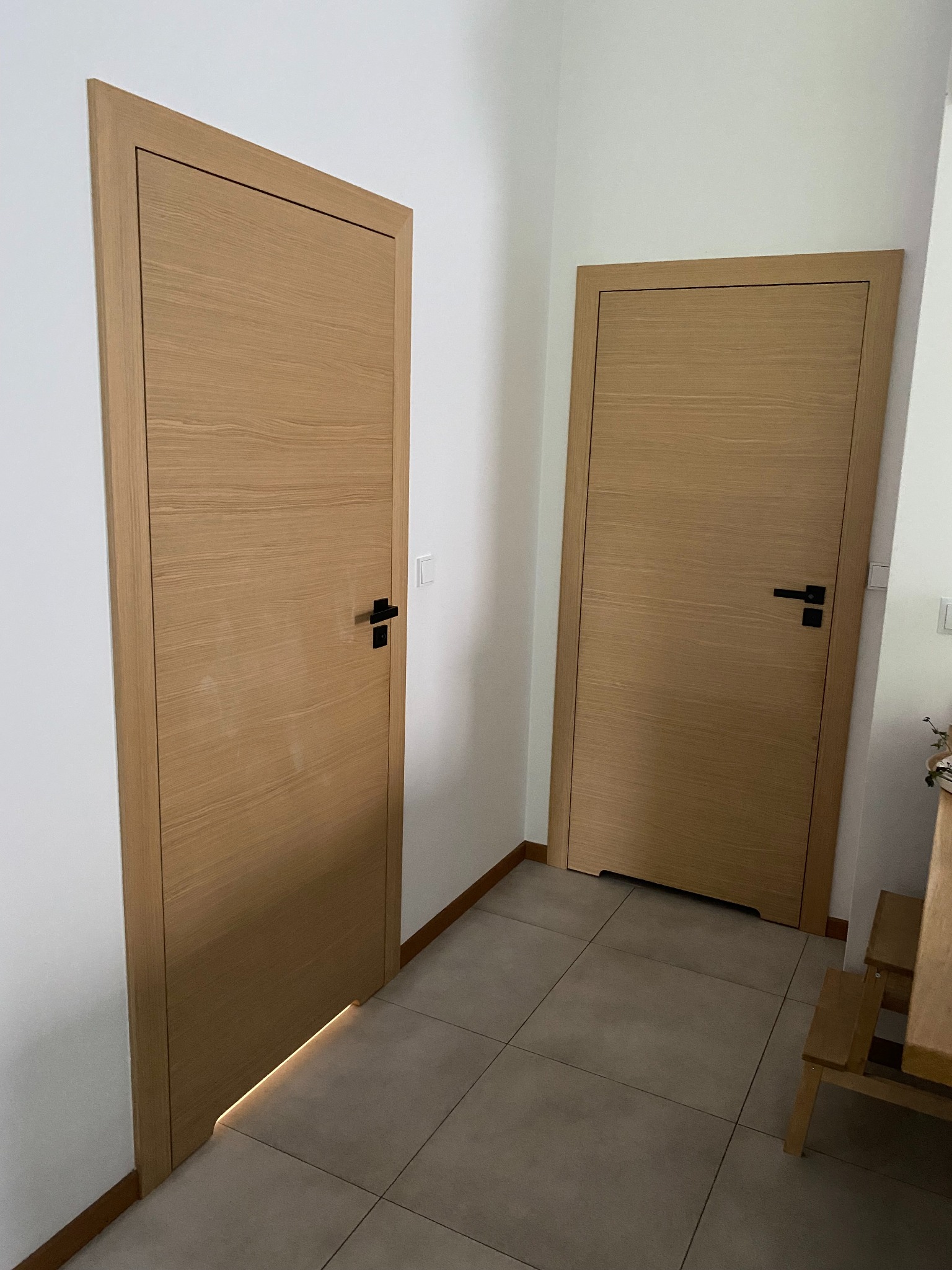 Dwa skrzydła drzwi wewnętrznych z naturalnego drewna, z czarnymi klamkami, zamontowane w jasnym korytarzu, z delikatnym podświetleniem dolnej krawędzi jednego skrzydła.