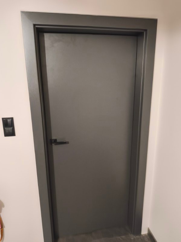 Szare, minimalistyczne drzwi wewnętrzne z czarną klamką i dopasowaną ościeżnicą, zamontowane w jasnym pomieszczeniu. Widoczny włącznik światła.