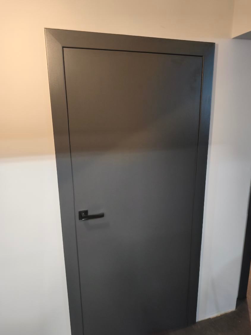 Nowoczesne, grafitowe drzwi wewnętrzne z minimalistyczną klamką, osadzone w białej ścianie. Prosta forma, stonowana kolorystyka, widoczny fragment podłogi.