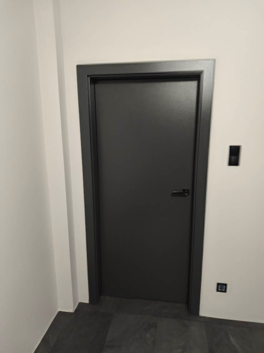 Matowe, grafitowe drzwi wewnętrzne z czarną klamką i ościeżnicą, na tle białej ściany z czarnymi włącznikami, na ciemnej podłodze. Minimalistyczny design.