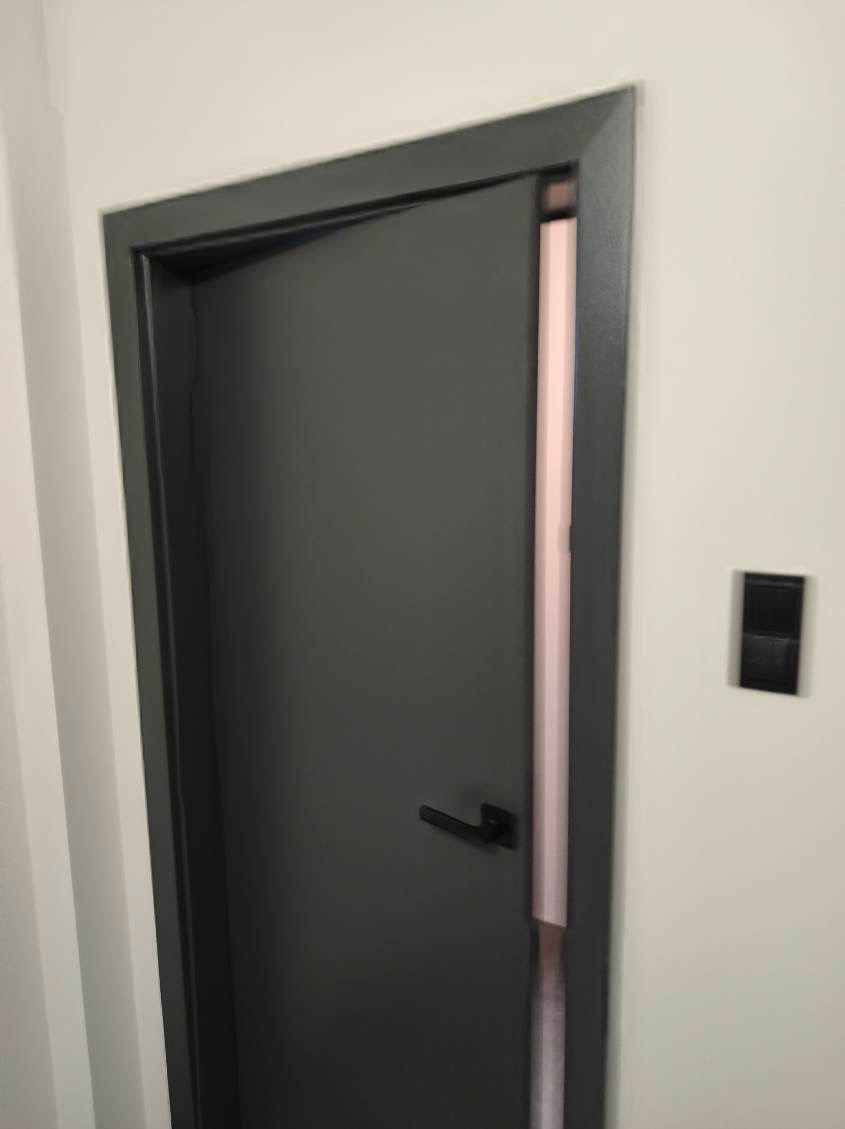 Grafitowe drzwi wewnętrzne z czarną klamką, częściowo otwarte, ukazujące różową uszczelkę. Minimalistyczny design, widoczny włącznik światła na białej ścianie.