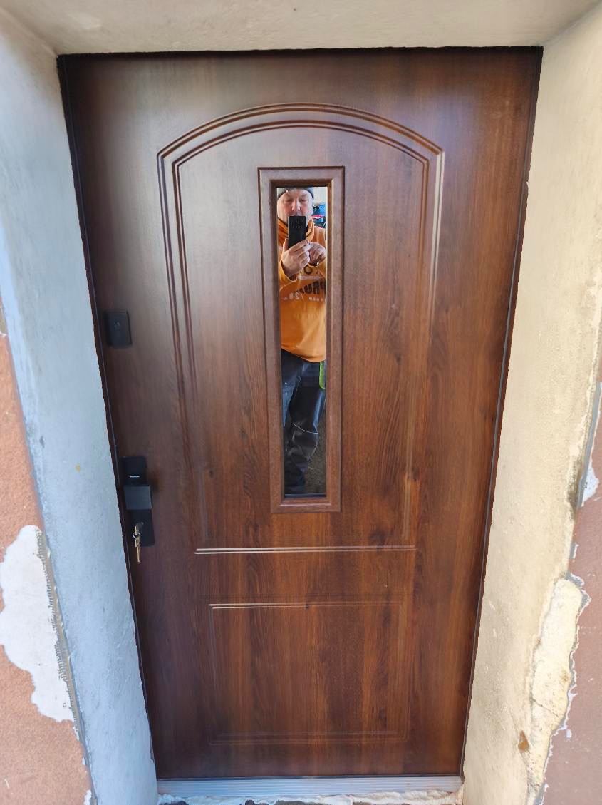 Brązowe drzwi zewnętrzne z szybą, widoczny zamek i klucz. W szybie odbija się osoba robiąca zdjęcie. Drzwi zamontowane w surowym otworze.