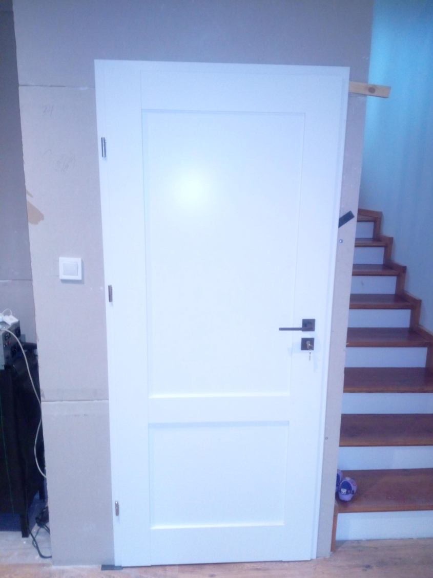 Białe, dwupanelowe drzwi wewnętrzne z czarną klamką i kluczem, zamontowane w surowym otworze, widoczne schody w tle. Minimalistyczny design.