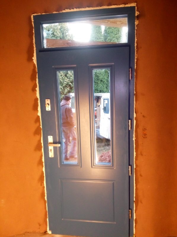 Niebieskie drzwi zewnętrzne z szybami, widoczne podczas montażu w surowym otworze. W szybach odbija się sylwetka osoby i zaparkowany biały samochód dostawczy.