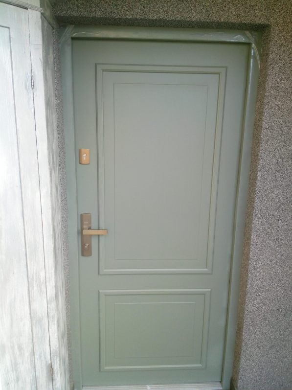 Zamknięte, jasne drzwi zewnętrzne z klamką i wizjerem, wkomponowane w szarą ścianę o ziarnistej fakturze. Widoczny fragment ościeżnicy i sąsiedniej ściany.
