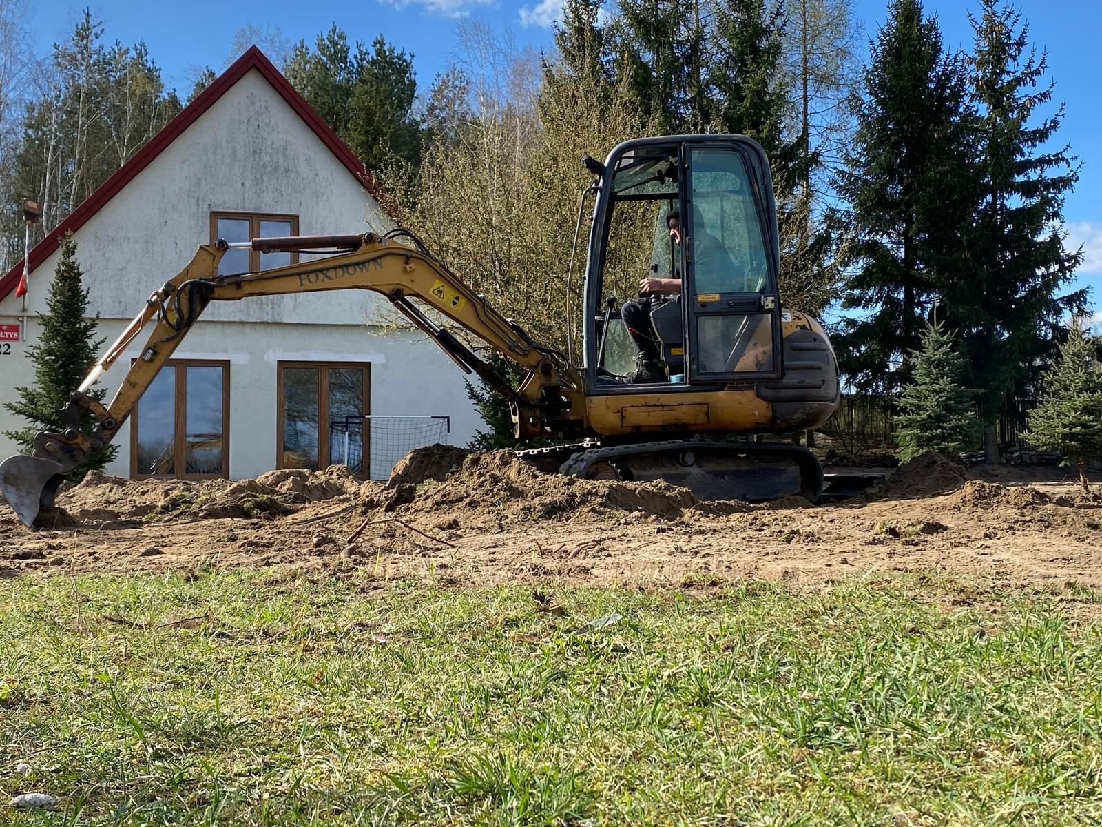 Żółta koparka Foxtown w trakcie prac ziemnych przed domem z brązowymi oknami i czerwonym dachem, w tle drzewa iglaste i błękitne niebo.