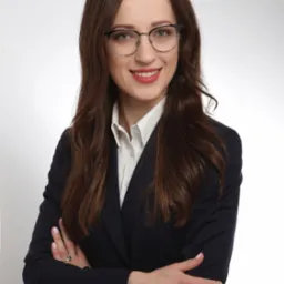 Portret uśmiechniętej brunetki w okularach i granatowym garniturze, skrzyżowane ręce, subtelny makijaż, delikatna biżuteria, jasne tło.