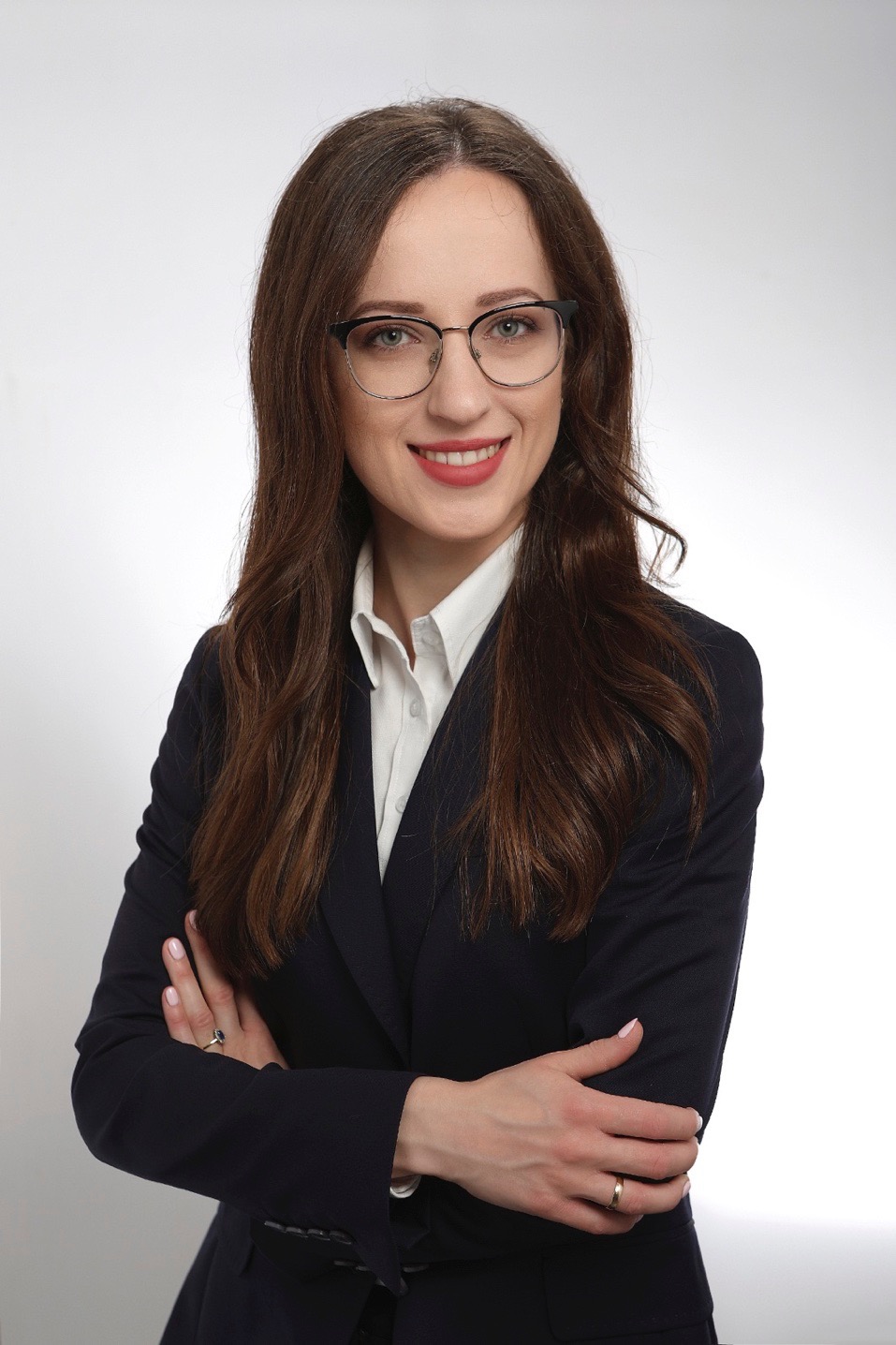 Portret uśmiechniętej brunetki w okularach i granatowym garniturze, skrzyżowane ręce, subtelny makijaż, delikatna biżuteria, jasne tło.