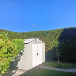 Metalowy domek ogrodowy na trawniku, otoczony wysokim, równo przyciętym żywopłotem i krzewami winorośli w słoneczny dzień. Czyste niebo.