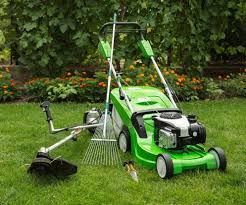 Zestaw narzędzi ogrodniczych: zielona kosiarka, podkaszarka, grabie i sekator na tle zielonego trawnika i krzewów. Sprzęt gotowy do użycia, w słoneczny dzień.