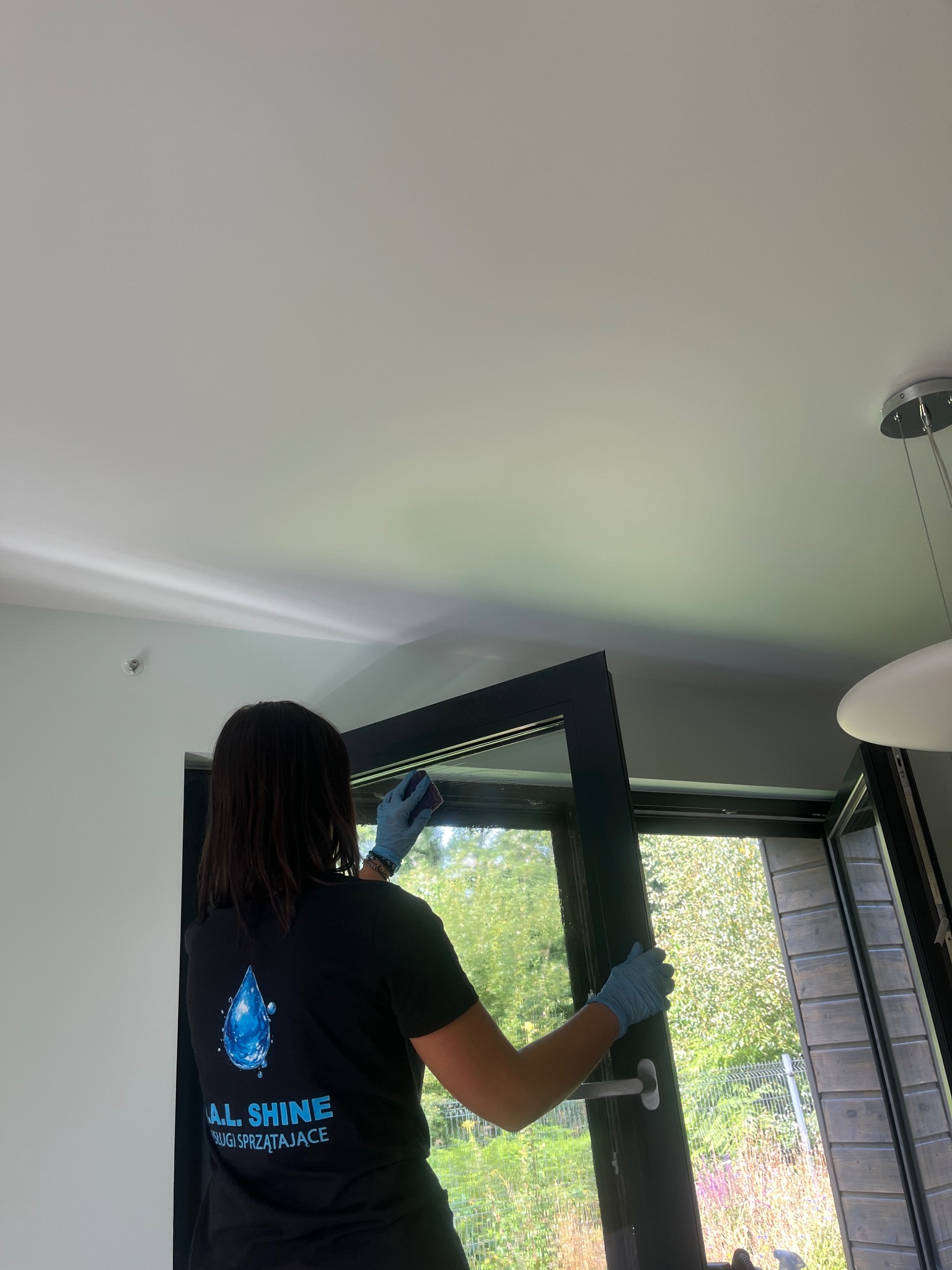 Osoba w niebieskich rękawicach czyści okno od wewnątrz, widoczny fragment ogrodu za oknem i logo firmy sprzątającej na koszulce pracownika.