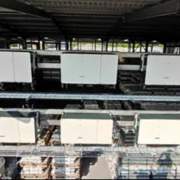 Rozbudowana instalacja fotowoltaiczna z białymi falownikami na regałach w hali przemysłowej, widok z góry, symetryczny układ i metalowa konstrukcja wsporcza.