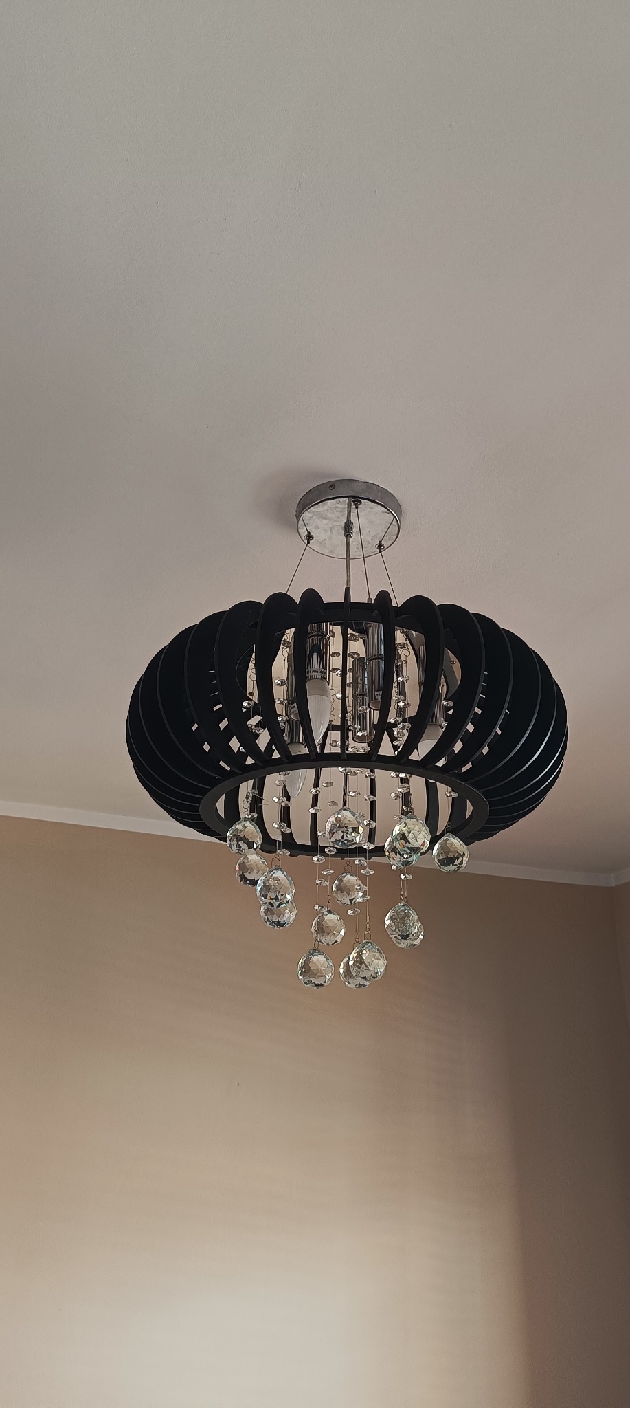 Czarny, okrągły żyrandol z kryształkami zwisającymi na tle beżowej ściany i białego sufitu. Widoczne żarówki i metalowe elementy mocujące.