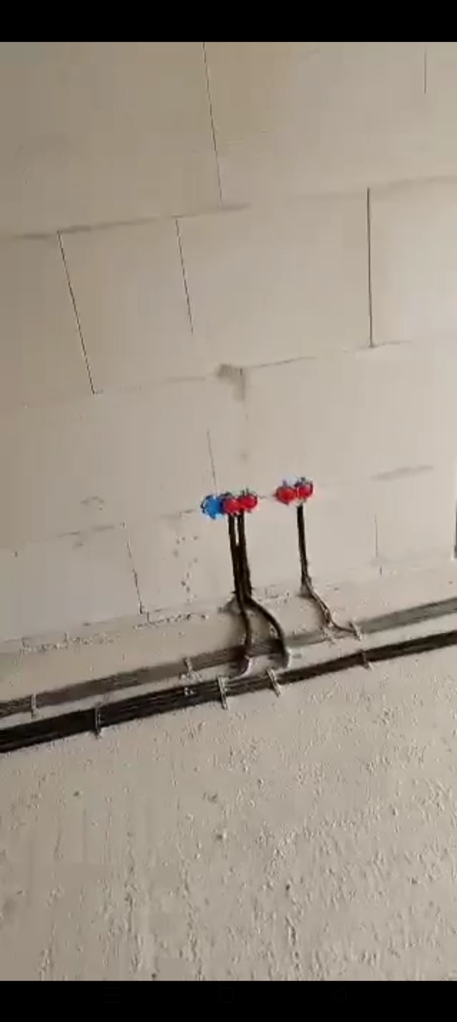 Fragment ściany z widocznymi, zamontowanymi przewodami elektrycznymi i rurami instalacji wodnej z czerwono-niebieskimi zaworami, przed dalszymi pracami wykończeniowymi.