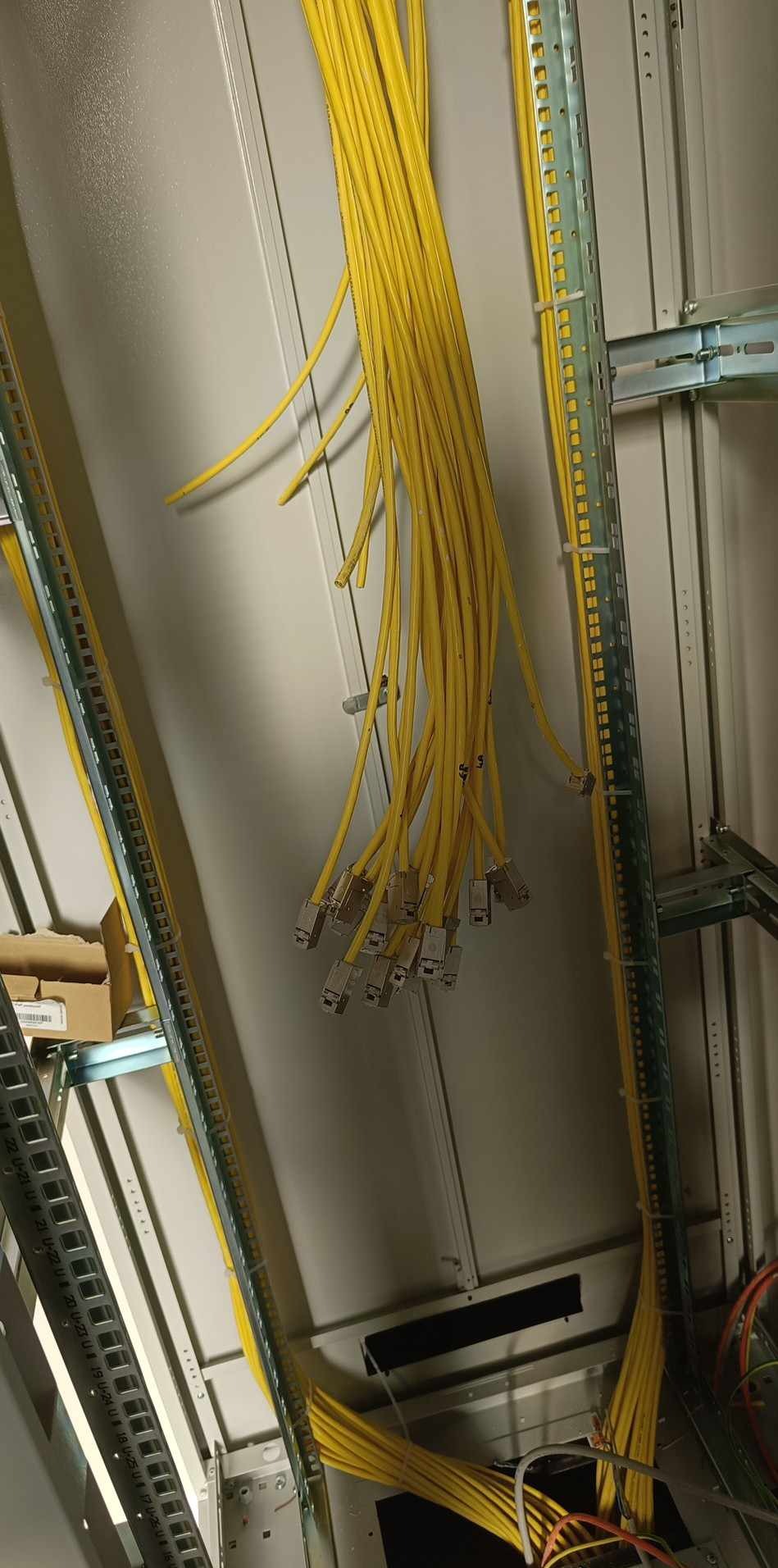 Żółte kable sieciowe z wtyczkami zwisają w szafie rack, uporządkowane za pomocą uchwytów. Widoczne metalowe szyny montażowe i fragmenty innych kabli.
