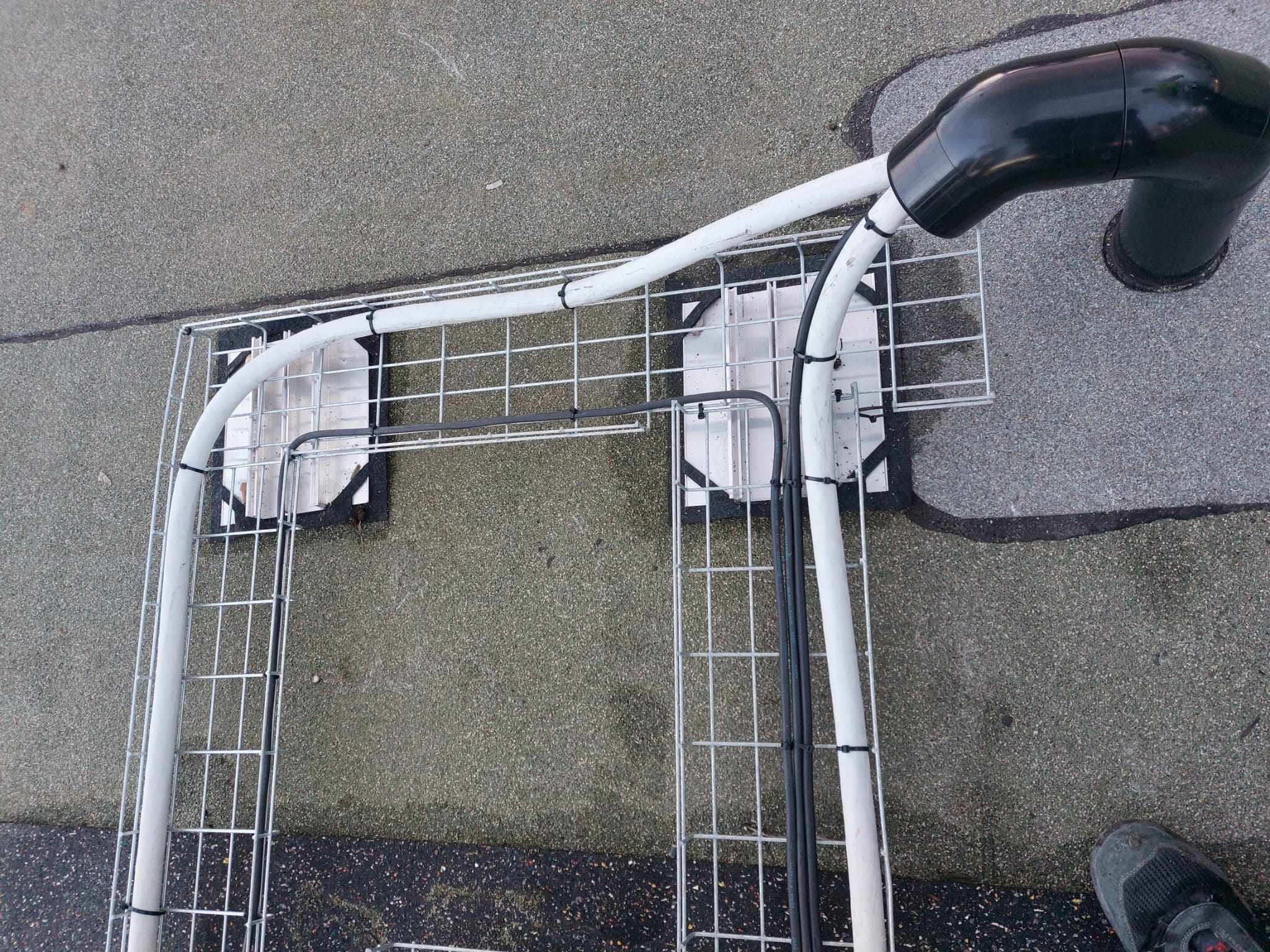 Klatka montażowa z przewodami elektrycznymi na dachu, widok z góry. Białe rury i czarne przewody przymocowane do metalowej konstrukcji. Fragment buta w prawym dolnym rogu.