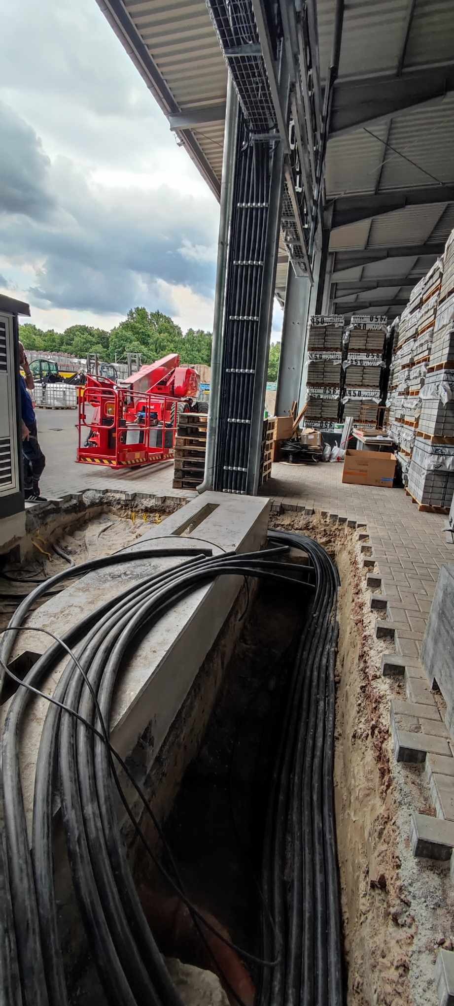 Kable w ziemi, prawdopodobnie instalacja elektryczna w hali. Widoczny wykop, ułożone kable, część konstrukcji hali i podnośnik koszowy w tle.