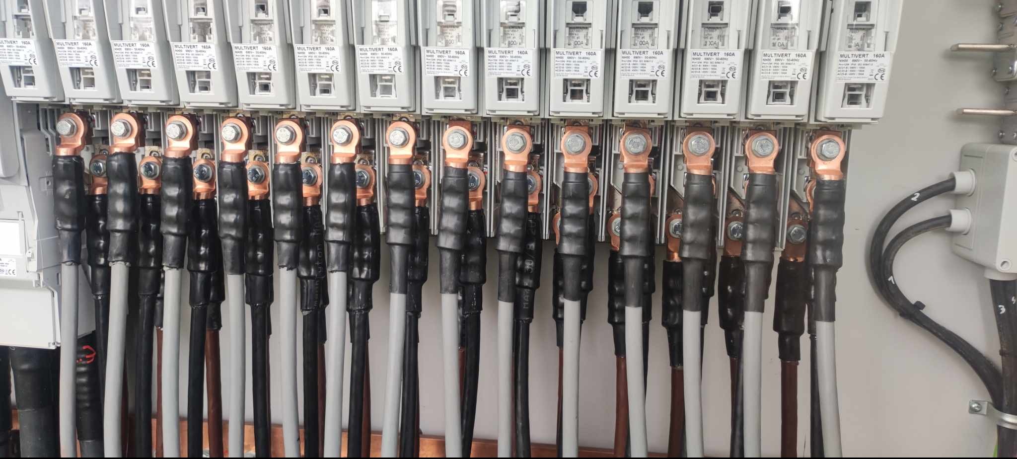 Szereg zabezpieczeń MULTIVERT 160A z podłączonymi przewodami w szarych i czarnych osłonach, zakończonych miedzianymi końcówkami. Widoczna skrzynka elektryczna.