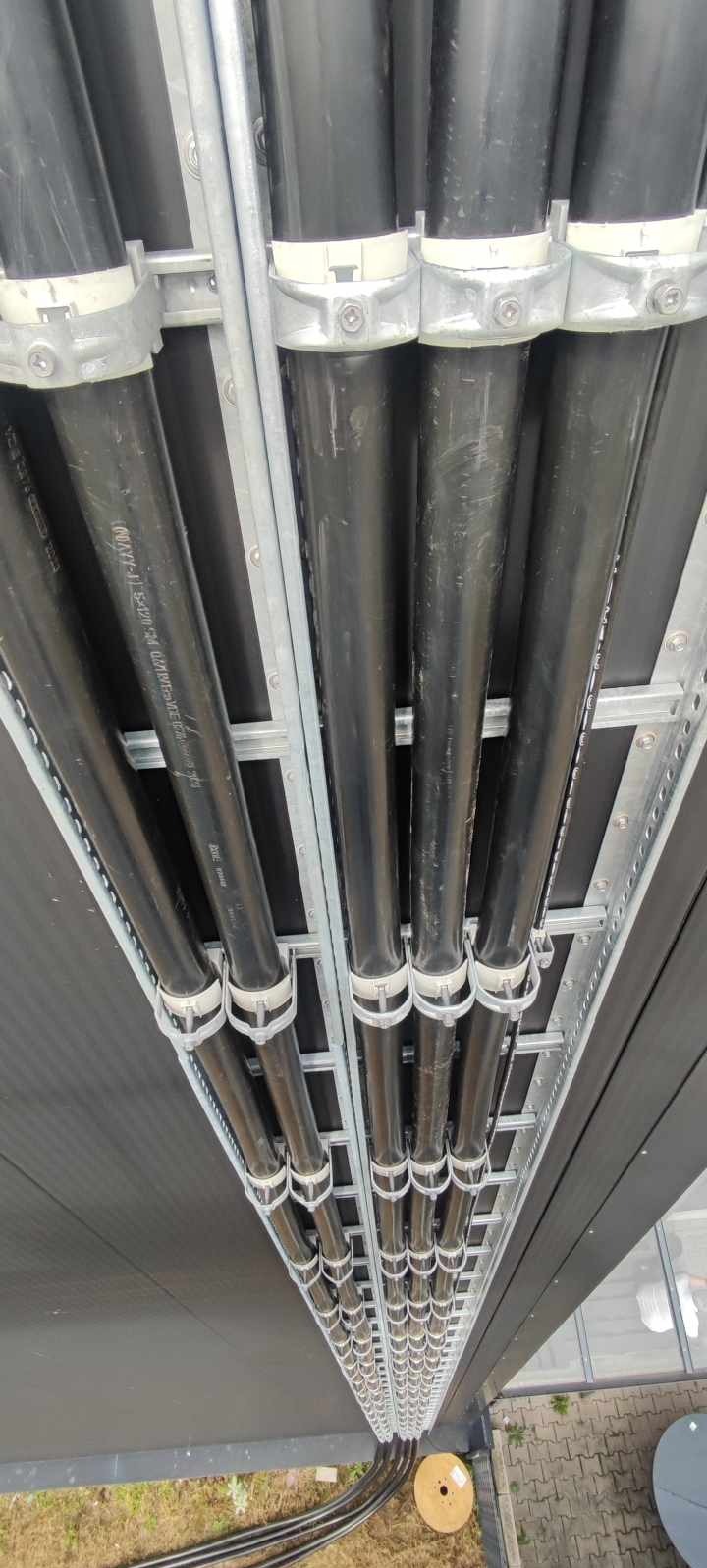 Rząd czarnych kabli elektrycznych zamocowanych na metalowej konstrukcji, widok z dołu pod kątem. Widoczne mocowania i fragmenty instalacji.