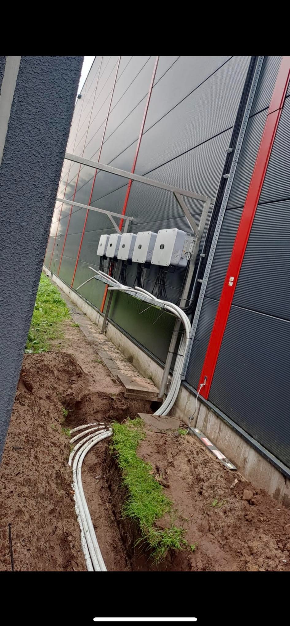 Montaż instalacji elektrycznej: białe kable w ziemi prowadzą do czterech szarych skrzynek na szarej ścianie budynku z czerwonymi akcentami.