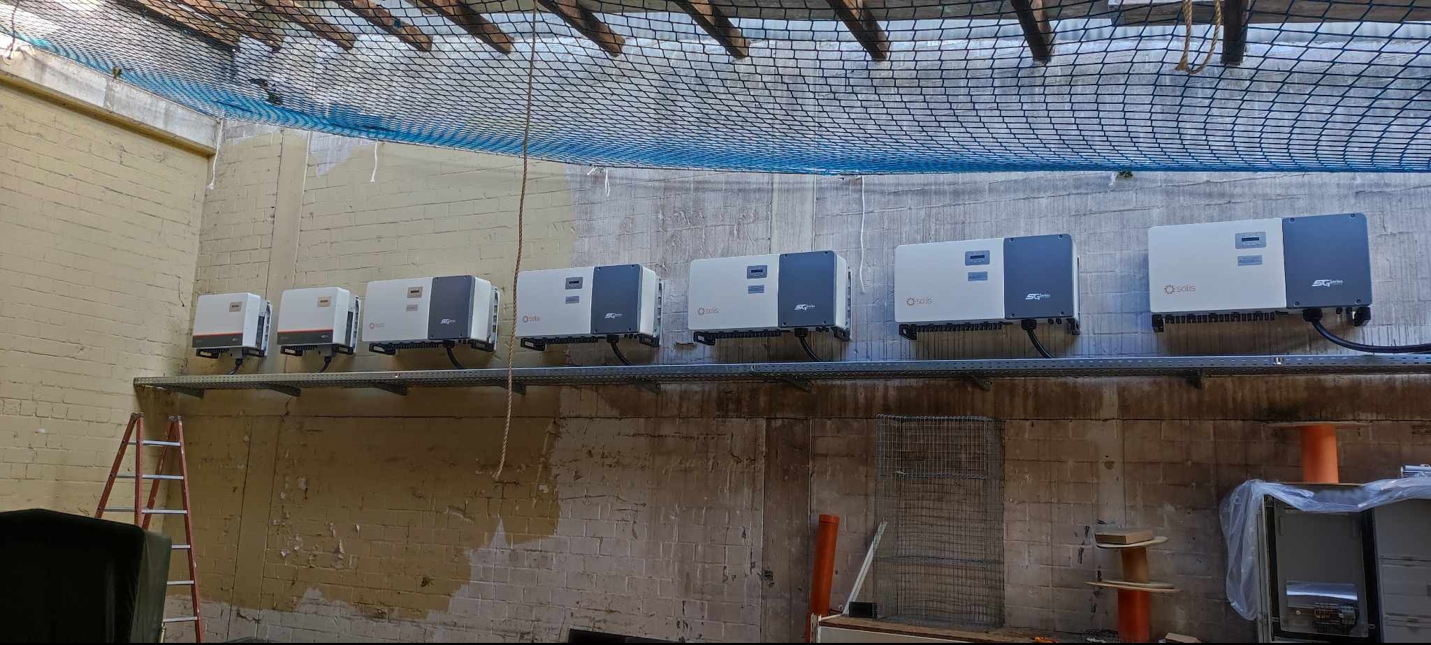 Rząd zamontowanych inwerterów Solis SG na metalowej szynie w surowym wnętrzu, zabezpieczonych siatką. Widoczna drabina i elementy instalacji.
