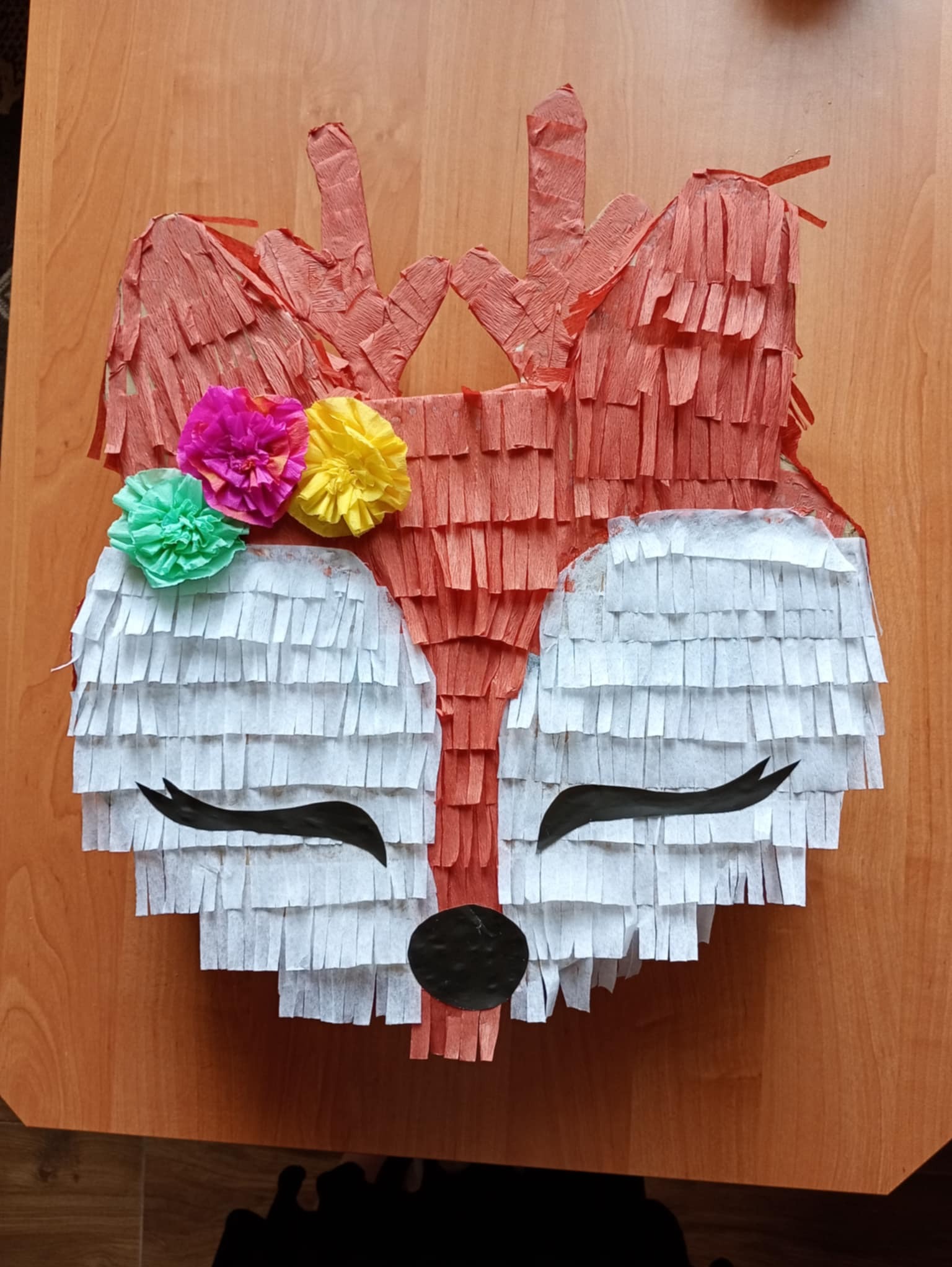 Ręcznie robiona piniata w kształcie liska, z krepy w odcieniach czerwieni i bieli, ozdobiona kolorowymi kwiatami, leży na drewnianym blacie.