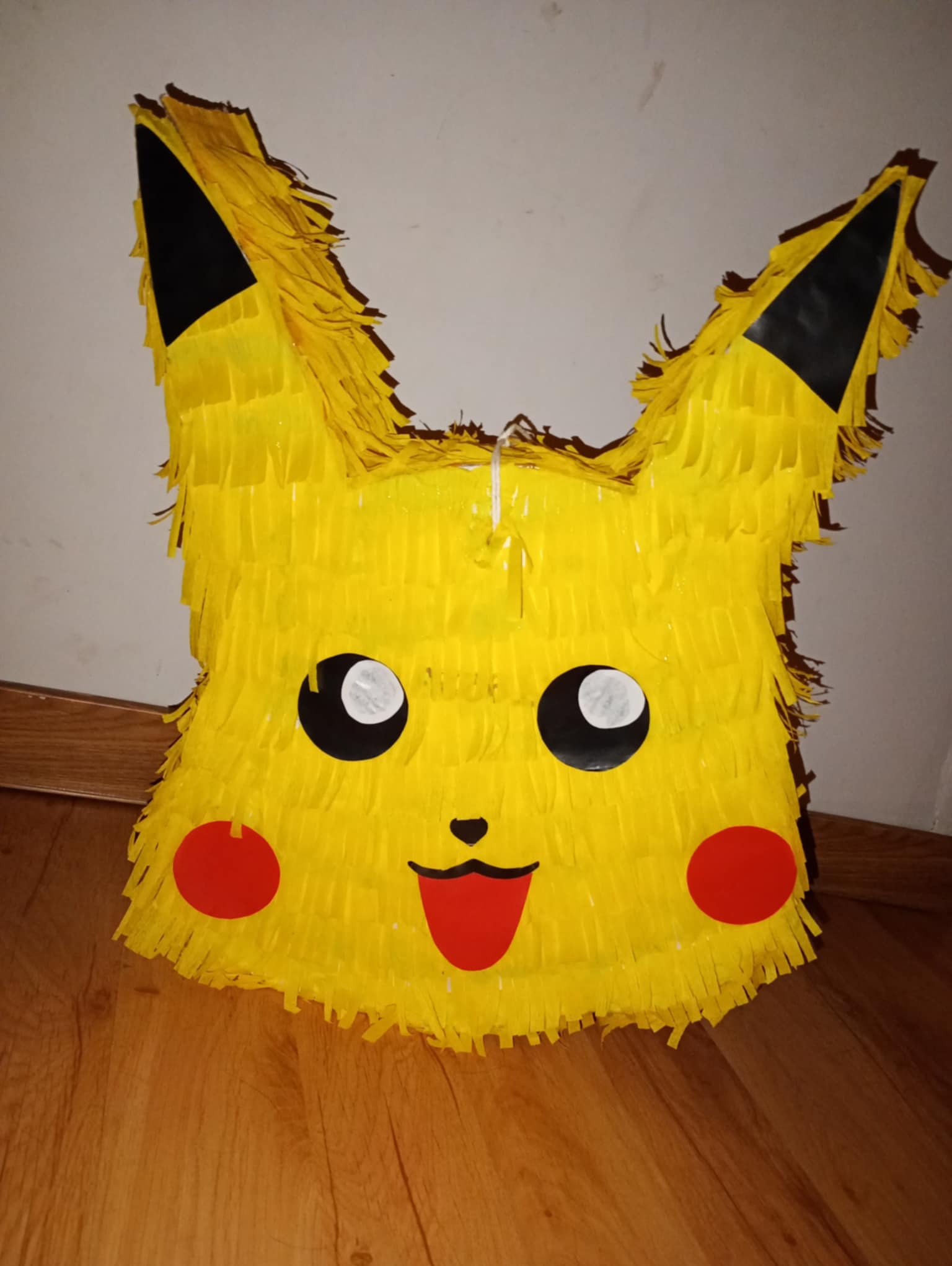 Żółta piniata w kształcie Pikachu z papierowymi frędzlami, czarnymi uszami i czerwonymi policzkami, leżąca na drewnianej podłodze. Idealna na imprezę dla dzieci.