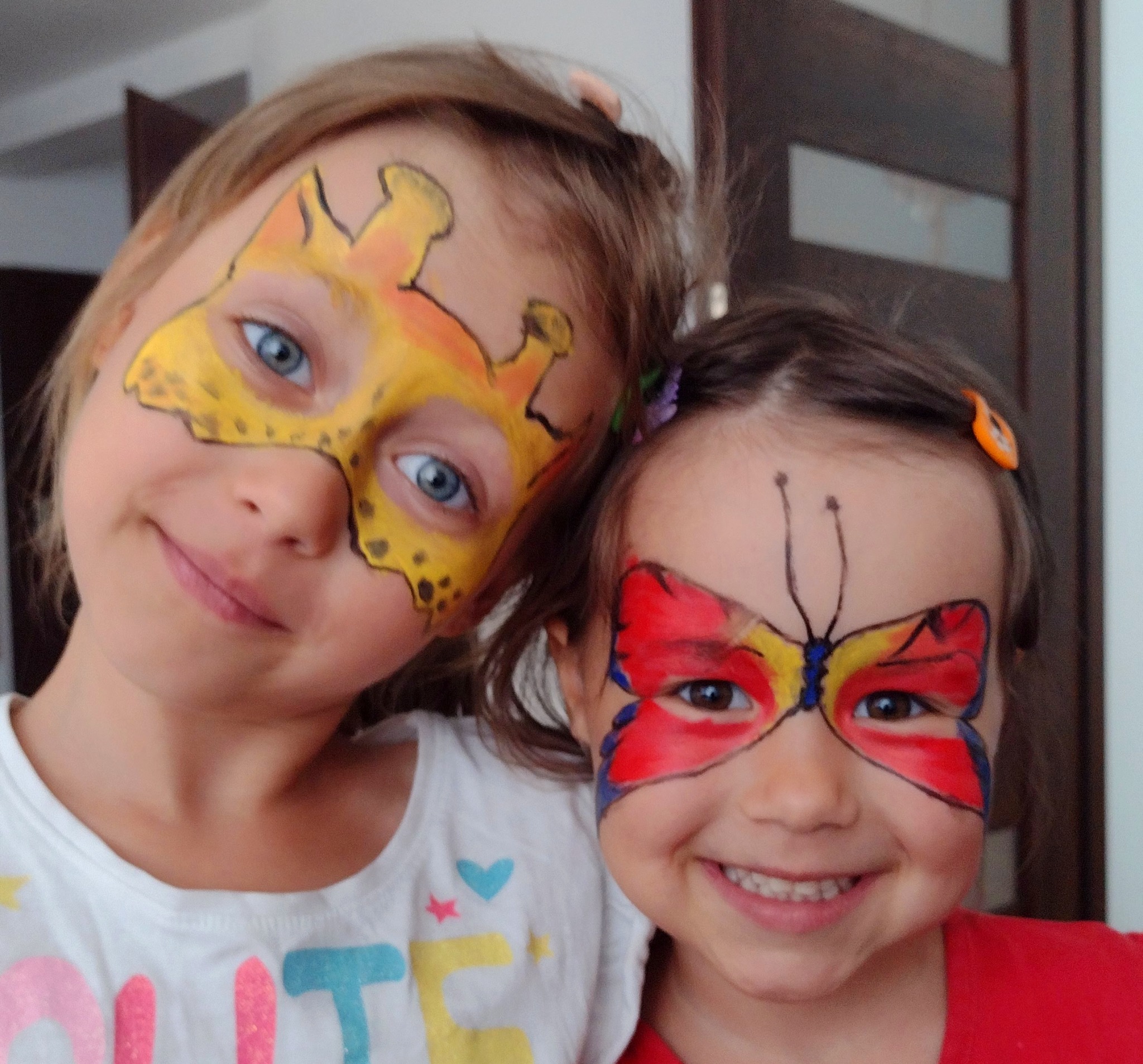 Dwie uśmiechnięte dziewczynki z pomalowanymi twarzami: jedna w motyw żyrafy, druga w motyw motyla. Wyraźne kolory, radosna atmosfera.