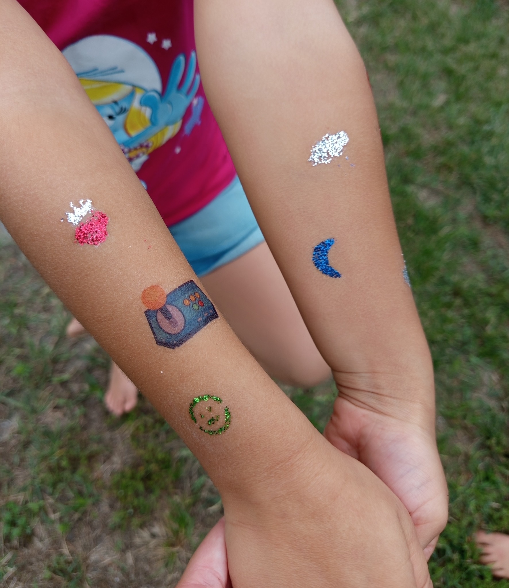 Dziecięce ramiona z brokatowymi tatuażami: joystick, uśmiechnięta buźka, księżyc i inne. T-shirt z wizerunkiem postaci z bajki w tle.