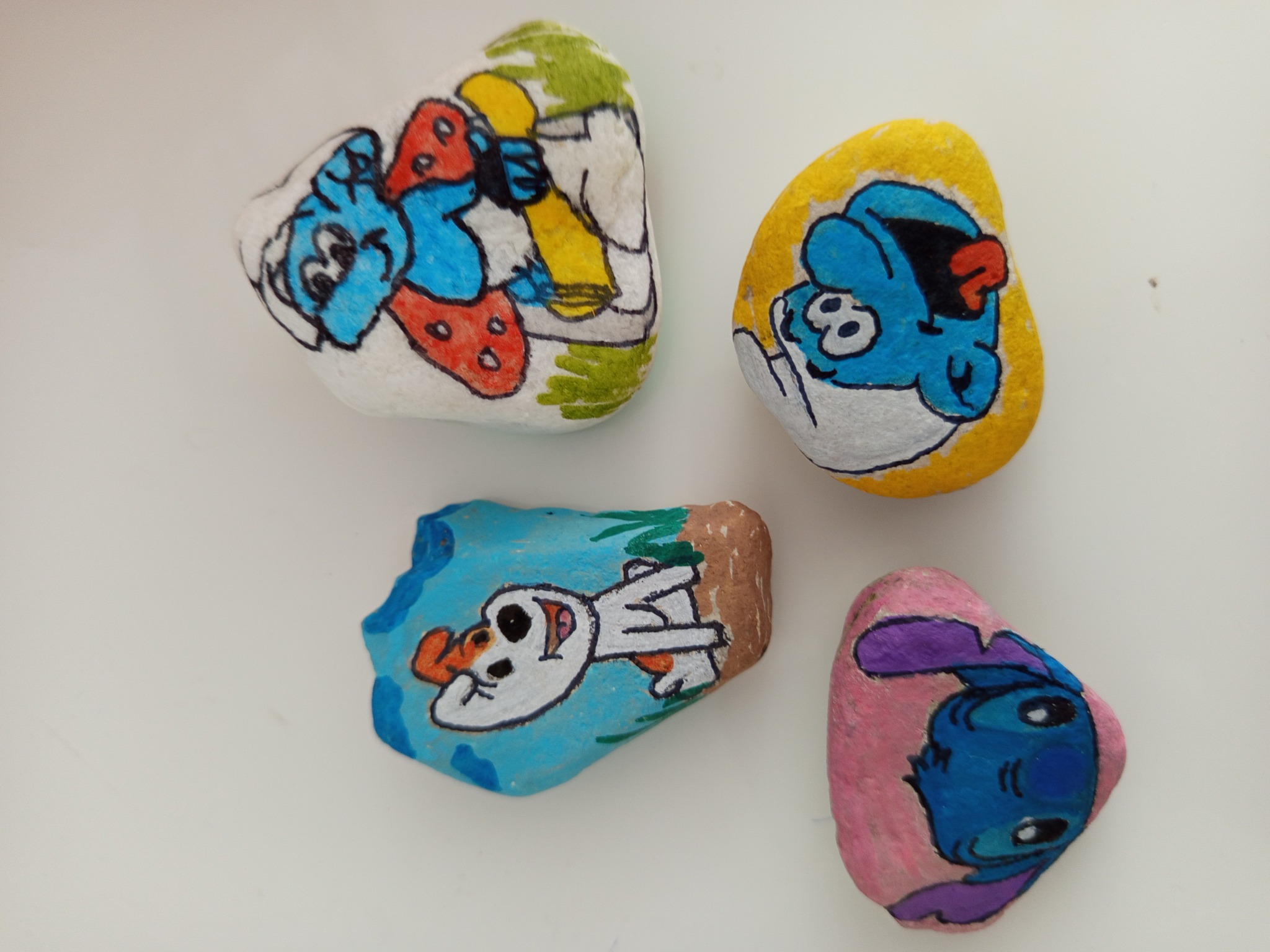 Ręcznie malowane kamienie z postaciami z bajek: Smerf, pies i Stitch, na białym tle. Idealne jako dekoracja na przyjęcie urodzinowe dla dzieci.
