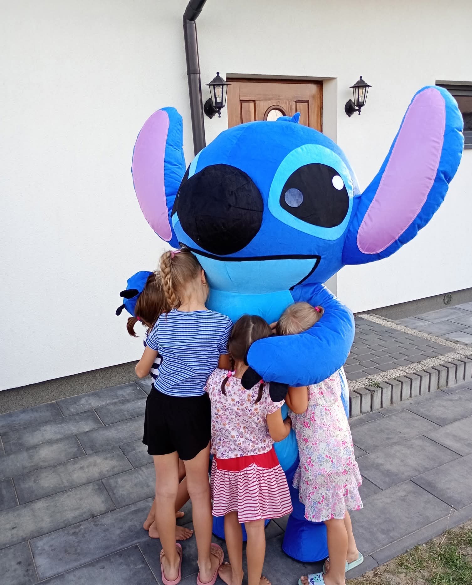 Dzieci tulące się do animatora w kostiumie Stitcha na tle białego budynku z brązowymi drzwiami i lampą. Kostium ma duże uszy i uśmiechniętą twarz.