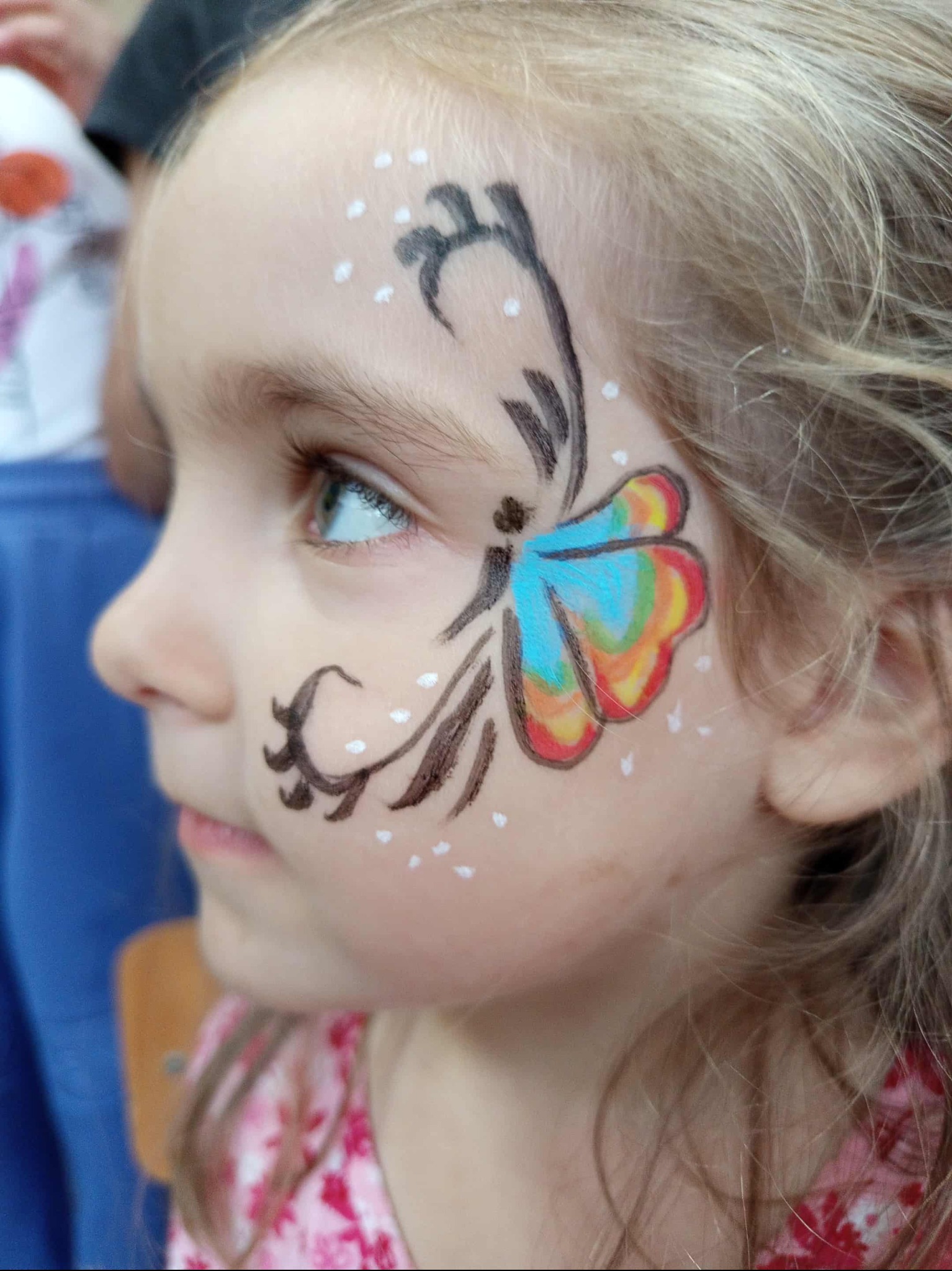 Profil dziewczynki z pomalowaną twarzą w motyla z czarnymi wzorami i białymi kropkami, kolorowe skrzydła, jasne włosy, delikatny uśmiech.