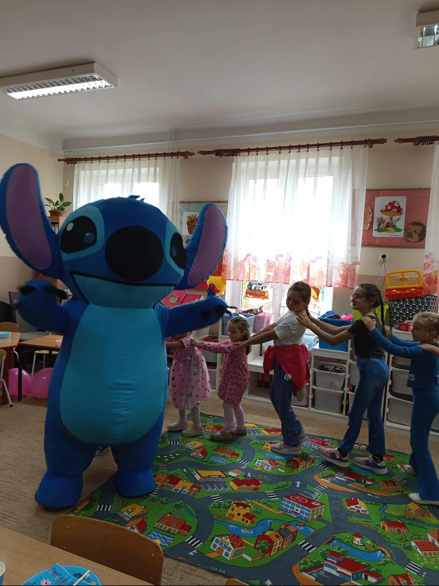 Animator Stitch prowadzi zabawę z dziećmi w Opole Lubelskie. Dzieci trzymają się za ręce, tworząc korowód. Kolorowy dywan z motywem miasta.