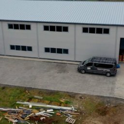 Haler Sp.z o.o. Sp.k. - Nowoczesna hala z szarej płyty warstwowej z bramą rolowaną, trzema autami na brukowanym parkingu i składowiskiem odpadów budowlanych. Ujęcie z drona.