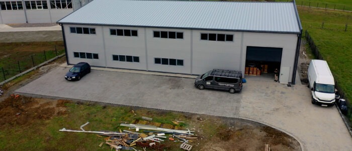 Nowoczesna hala z szarej płyty warstwowej z bramą rolowaną, trzema autami na brukowanym parkingu i składowiskiem odpadów budowlanych. Ujęcie z drona.