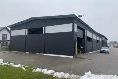 Nowoczesna hala z płyt warstwowych w kolorze grafitowym, z widocznymi poziomymi pasami w kolorze srebrnym. Częściowo otwarte wrota, fragmenty śniegu przy budynku.