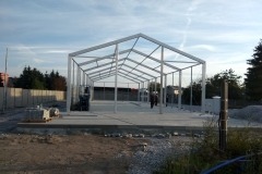 Stalowa konstrukcja hali w trakcie budowy, widok z zewnątrz. Szary betonowy fundament, białe profile, drzewa w tle i fragment betonowego ogrodzenia.