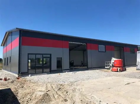 Nowoczesna hala stalowa z grafitowo-czerwonymi panelami, częściowo otwarta, z widocznym wnętrzem i podnośnikiem koszowym na zewnątrz.