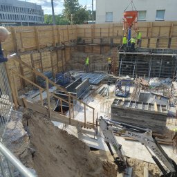 Pitbul AMKD Oleksandr Pitel - Wykop pod budynek z widocznym zbrojeniem i szalunkami. Robotnicy w kaskach i kamizelkach wylewają beton za pomocą pompy. Mini koparka na pierwszym planie.