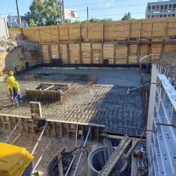 Pitbul AMKD Oleksandr Pitel - Zalewanie fundamentów na budowie. Widoczni pracownicy w żółtych kamizelkach, zbrojenie i szalunki. Ujęcie z góry na wykop z zabezpieczonymi ścianami.