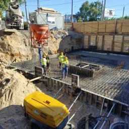 Pitbul AMKD Oleksandr Pitel - Wylewanie betonu na zbrojenie fundamentów w Rumi. Na budowie widoczni pracownicy w kaskach i żółtych kamizelkach. Na pierwszym planie sprężarka.