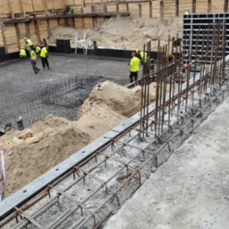 Zbrojenie fundamentów w trakcie budowy. Widoczne stalowe pręty, szalunki i pracownicy w żółtych kamizelkach. Na drugim planie budynek w budowie.