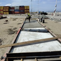 Pitbul AMKD Oleksandr Pitel - Wykonywanie betonowych fundamentów w szalunkach na placu budowy. W tle widoczne kontenery i pracownik w kamizelce odblaskowej. Ujęcie z góry.