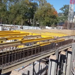 Wysoki kąt: Zbrojenie pionowe i poziome, żółte belki wspierające na placu budowy. W tle budynek i drzewa w jesiennych barwach. Częściowo widoczny żuraw.