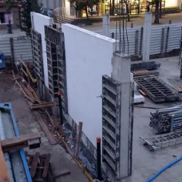 Budowa ściany z bloczków betonowych z szalunkiem i wystającymi prętami zbrojeniowymi. Widoczne elementy konstrukcyjne i narzędzia budowlane na placu budowy.