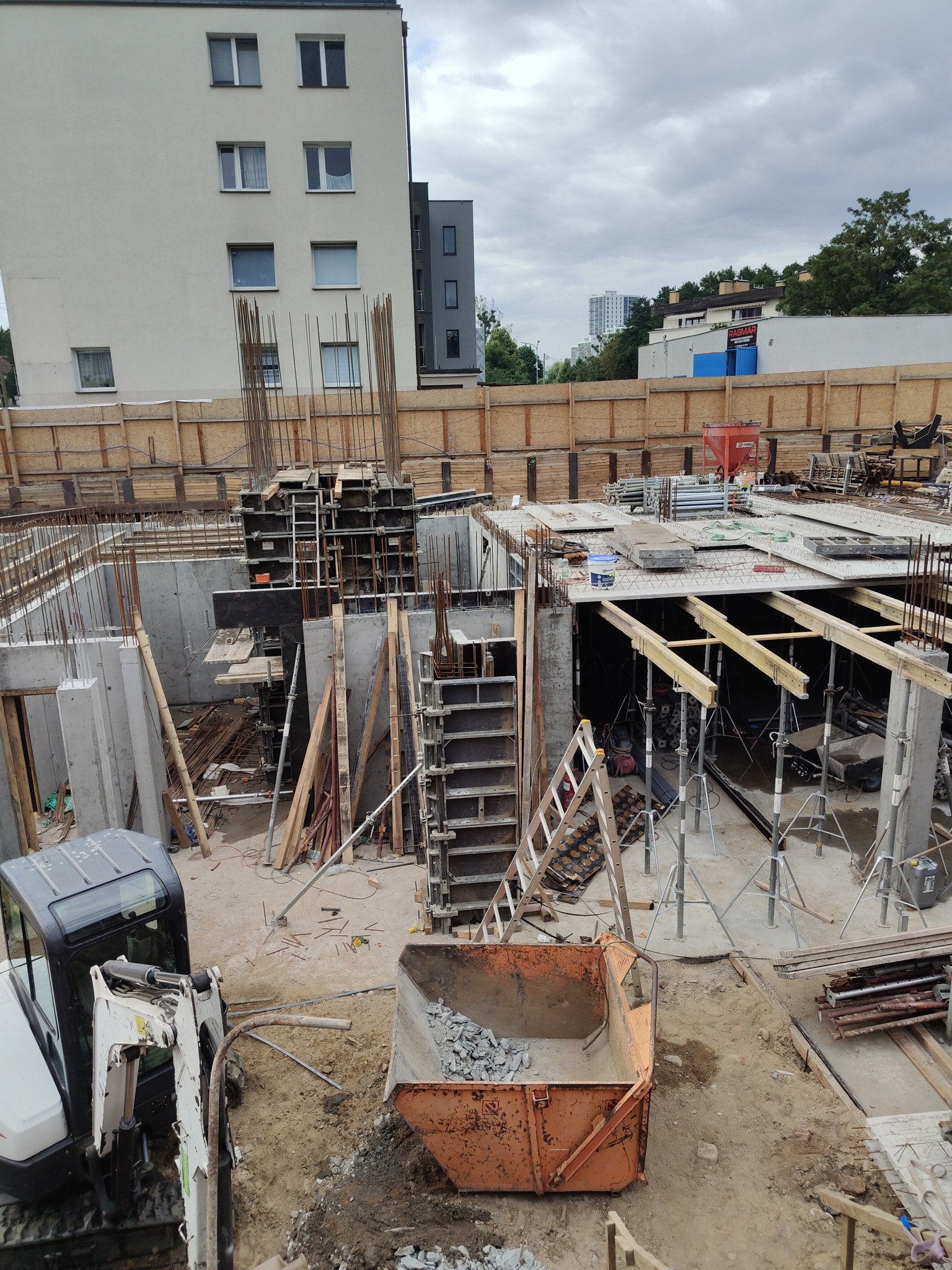 Wykop fundamentowy z częściowo wykonanym zbrojeniem i deskowaniem. Widoczny minikoparka i kontener na gruz. W tle zabudowa miejska i pochmurne niebo.