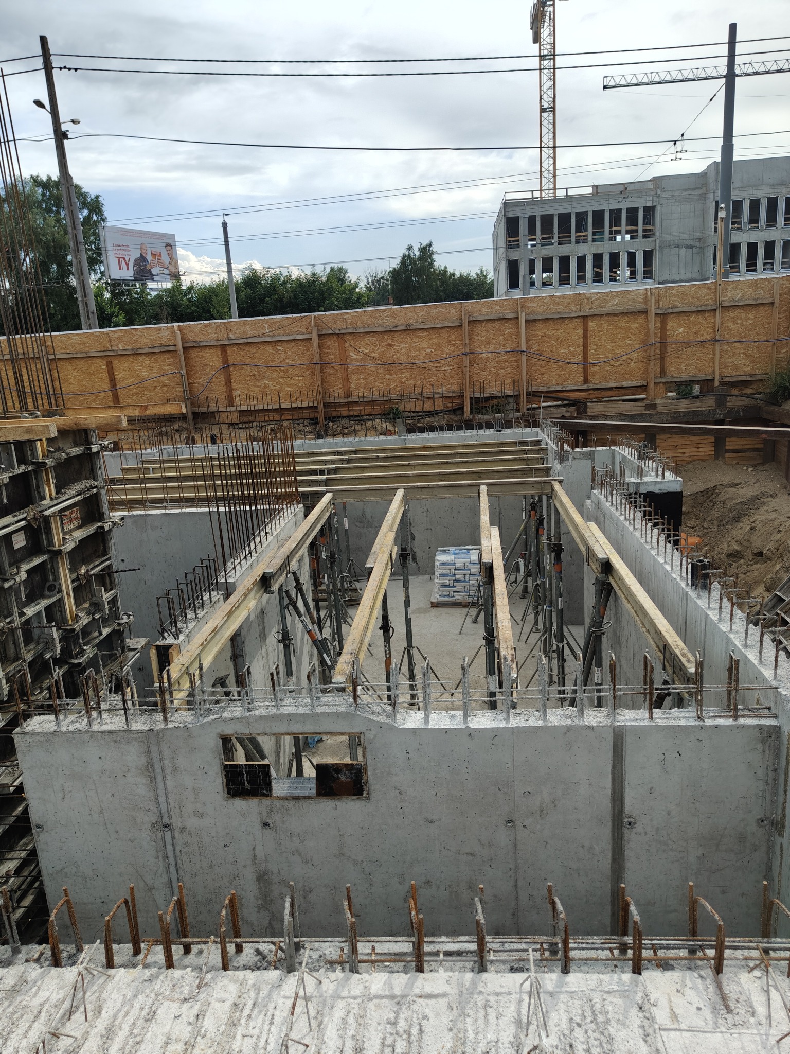 Betonowy fundament budynku w trakcie budowy, z widocznym zbrojeniem i szalunkiem. W tle widać częściowo ukończony budynek i dźwig budowlany.