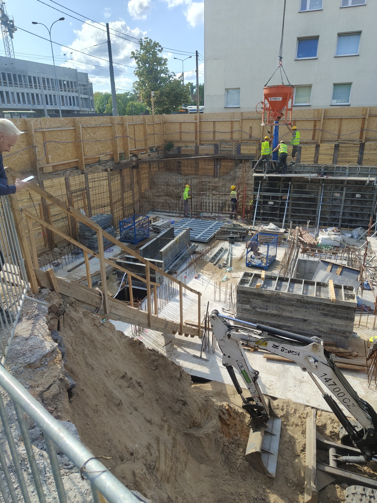 Wykop pod budynek z widocznym zbrojeniem i szalunkami. Robotnicy w kaskach i kamizelkach wylewają beton za pomocą pompy. Mini koparka na pierwszym planie.